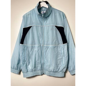 Puede incluir: Chaqueta azul claro con cremallera y detalles en negro y blanco. La chaqueta tiene cuello alto, cremallera frontal y dos bolsillos delanteros. Las mangas y la cintura tienen bandas elásticas. La chaqueta está colgada de una percha blanca.