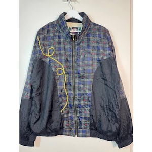 Puede incluir: Chaqueta vintage con un estampado de cuadros azules, verdes y rojos, con paneles negros en las mangas y un diseño decorativo amarillo. La chaqueta tiene una cremallera completa y está colgada de una percha blanca.