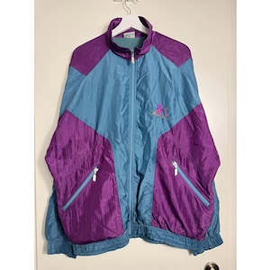 Op de afbeelding: Een vintage trainingsjack in een colorblock-ontwerp. De jas heeft een blauwgroene body met paarse accenten op de schouders en de onderkant van de voorkant. Het heeft een volledige ritssluiting, twee voorzakken en een klein geborduurd logo op de borst.