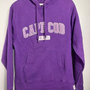 Könnte beinhalten: Lila Kapuzenpullover mit dem Text "CAPE COD" in weißer und hellvioletter Schrift. Das Wort "MASS" steht darunter. Der Pullover hat eine Vordertasche und eine Kapuze mit Kordelzug. Er hängt an einem weißen Kleiderbügel.