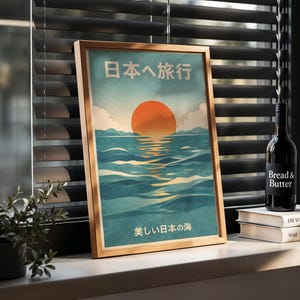 May include: A framed print with Japanese text and a vintage travel poster design. The artwork depicts a vibrant orange sun setting over blue ocean waves. A bottle of wine and books are on the windowsill.
