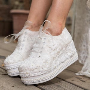 May include: White lace platform sneakers with floral and pearl embellishments. The sneakers have a thick sole and are tied with sheer ribbons. The design features intricate lace detailing and a classic sneaker silhouette.
