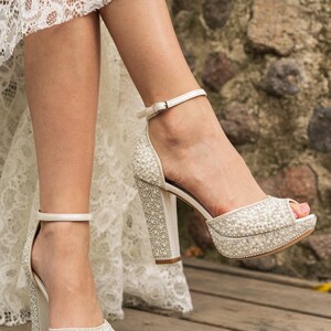 Pu&ograve; includere: Sandali bianchi con tacco alto e plateau, impreziositi da perle. Le scarpe a punta aperta hanno un tacco spesso e un cinturino alla caviglia. Le scarpe sono su una superficie di legno e lo sfondo &egrave; un muro di pietra.