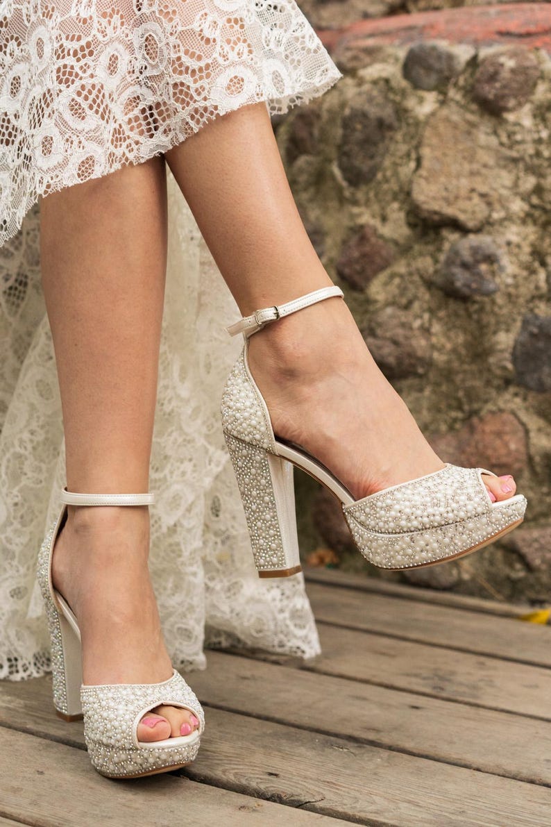 Pu&ograve; includere: Sandali bianchi con tacco alto e plateau, impreziositi da perle. Le scarpe a punta aperta hanno un tacco spesso e un cinturino alla caviglia. Le scarpe sono su una superficie di legno e lo sfondo &egrave; un muro di pietra.