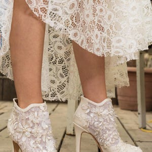 Puede incluir: Botines de encaje y sat&eacute;n color marfil con tacones altos. Las botas presentan adornos florales y con cuentas. La imagen muestra las botas sobre una superficie de madera, con un vestido de encaje visible.