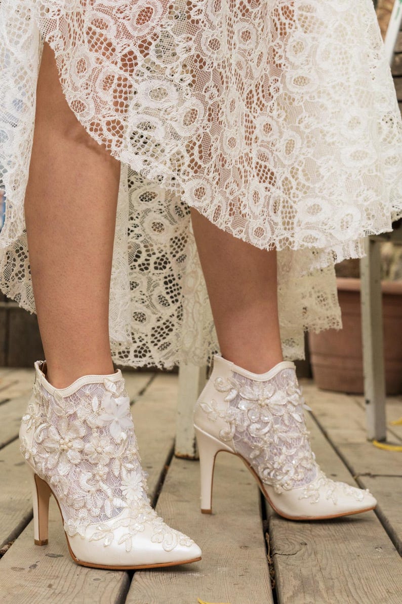 Puede incluir: Botines de encaje y sat&eacute;n color marfil con tacones altos. Las botas presentan adornos florales y con cuentas. La imagen muestra las botas sobre una superficie de madera, con un vestido de encaje visible.