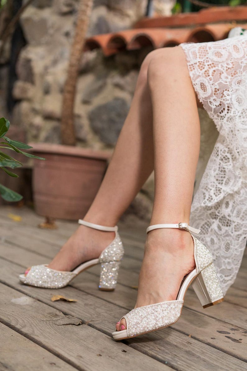May include: Sparkling white bridal heels with a peep-toe design. The heels feature a thick, block heel and an ankle strap with a buckle. The shoes are covered in small, shimmering embellishments, perfect for a wedding or special occasion.