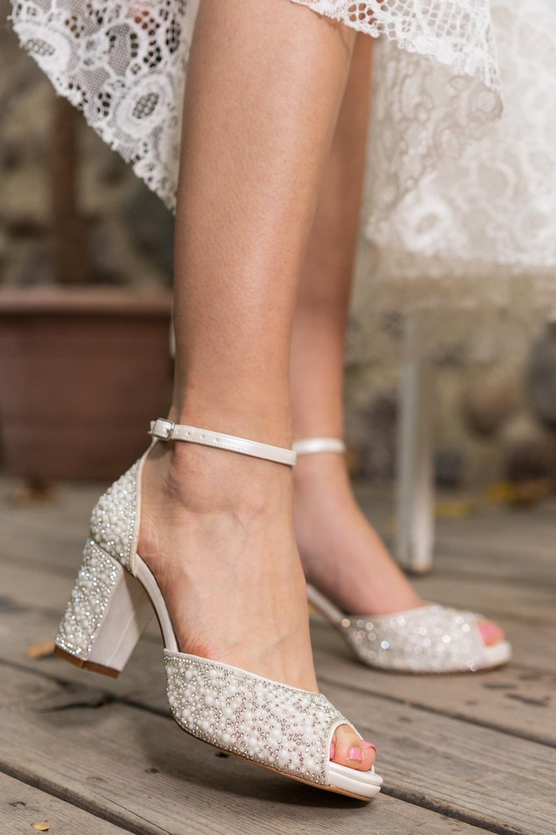 May include: White bridal heels with a block heel and ankle strap. The open-toe shoes are embellished with small, shiny beads. The heels are approximately 3 inches high.
