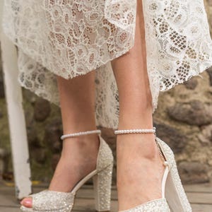 Pu&ograve; includere: Scintillanti tacchi alti bianchi con plateau con braccialetto alla caviglia di perle. Le scarpe a punta aperta sono ricoperte di decorazioni scintillanti e hanno un tacco grosso. &Egrave; visibile anche un abito di pizzo bianco.