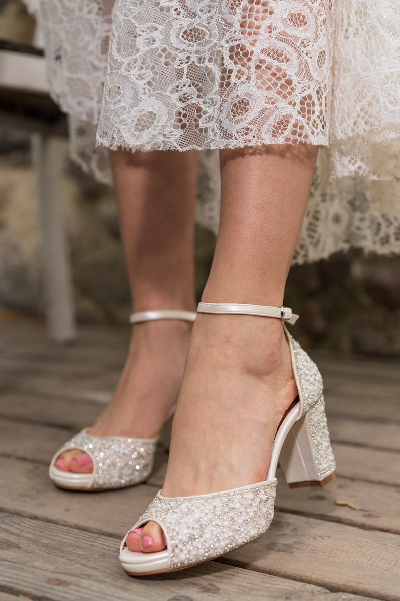 May include: White bridal peep-toe heels with pearl and rhinestone embellishments. The block heels have a matching pearl and rhinestone design. The shoes have a delicate ankle strap and are worn with a white lace dress.