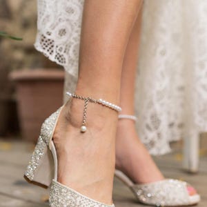 May include: Ivory peep-toe heels adorned with sparkling embellishments and a pearl ankle bracelet. The block heels and open-toe design create a classic bridal look. The shoes are set on a wooden surface.
