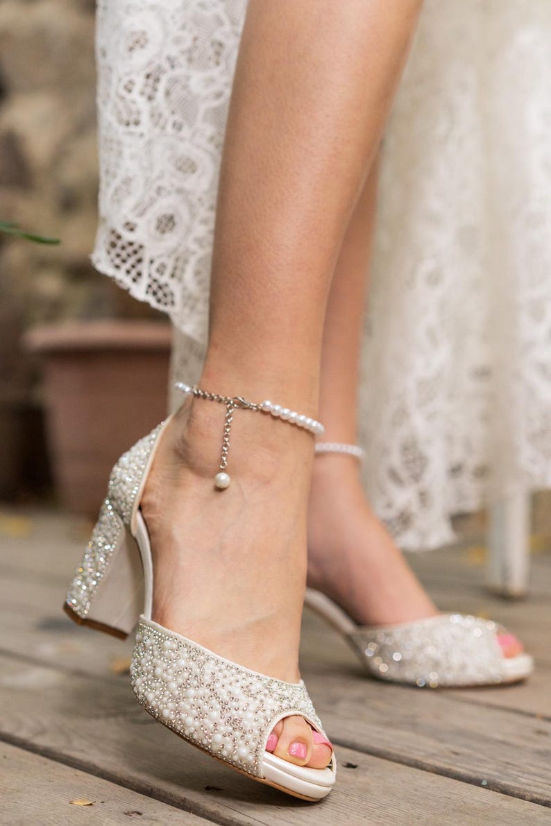 May include: Ivory peep-toe heels adorned with sparkling embellishments and a pearl ankle bracelet. The block heels and open-toe design create a classic bridal look. The shoes are set on a wooden surface.