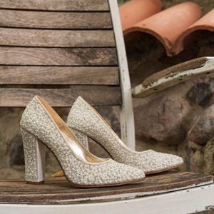 May include: A pair of ivory-colored high heels with a pearl-like embellishment. The heels have a chunky design and are set on a wooden chair with a distressed white paint finish. The background includes stone and terracotta elements.