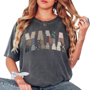 May include: Dark gray t-shirt with the word "MAMA" in patchwork letters. Each letter is composed of different fabric patterns, including floral, plaid, and geometric designs. The shirt has a vintage, worn look.