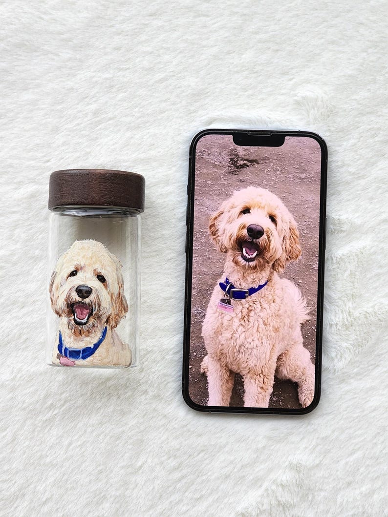 Puede incluir: Un tarro de cristal transparente con tapa de madera y un retrato pintado de un perro Goldendoodle con collar azul. Un smartphone muestra una imagen similar del perro. El perro tiene pelaje marr&oacute;n claro y est&aacute; sonriendo.