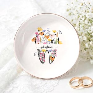 Op de afbeelding: Een witte keramische ringschaal met een gouden rand, versierd met een bloemen tand ontwerp en de naam "Sebastiane". Twee gouden trouwringen liggen naast de schaal. De schaal staat op een wit kanten oppervlak.