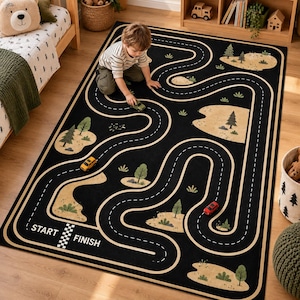 Puede incluir: Una alfombra rectangular negra con un patr&oacute;n de carretera sinuosa y marcas de carril blancas discontinuas. El dise&ntilde;o incluye parches de paisaje beige con &aacute;rboles y arbustos verdes. El texto START y FINISH est&aacute; impreso en blanco.