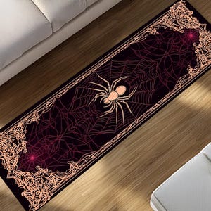 Puede incluir: Una alfombra rectangular con temática de Halloween con un diseño de araña y telaraña. La alfombra tiene un fondo negro con un patrón de telaraña en tonos rojos. Una araña grande está en el centro y el borde es de color melocotón claro.