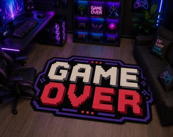 May include: A rectangular area rug featuring a pixelated design with the text "GAME OVER" in large white and red block letters. The rug has a black background with a purple border and decorative pixelated corner accents, mimicking a retro video game.