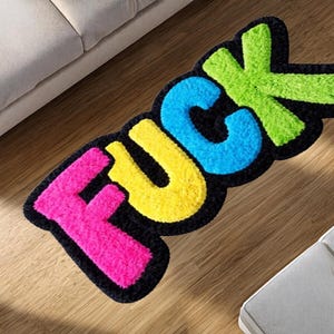 May include: A rectangular rug featuring the word "FUCK" in vibrant colours. The letters are pink, yellow, blue, and green, with a black outline. The rug is positioned on a wooden floor.