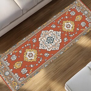 Puede incluir: Una alfombra rectangular con un diseño floral tradicional. La alfombra presenta un fondo naranja quemado con patrones florales crema, amarillos y azul marino. La alfombra está colocada sobre un suelo de madera, cerca de un sofá y una silla.