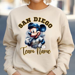 May include: Cream-colored sweatshirt featuring a cartoon Mickey Mouse in a baseball uniform. The design includes the text "SAN DIEGO" above Mickey and "Team Name" below. Mickey is holding a baseball bat and a baseball.