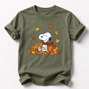 May include: An olive green short-sleeved t-shirt featuring a graphic of the cartoon character Snoopy wearing a red and black plaid scarf, sitting in a pile of colorful autumn leaves with additional leaves falling around the character.