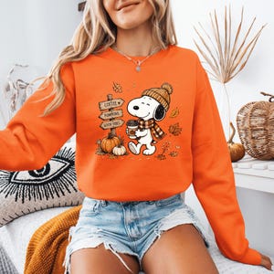 May include: An orange long-sleeve sweatshirt featuring a graphic of a cartoon dog wearing a knit hat and scarf, holding a coffee cup. Next to the dog is a wooden signpost with the text COFFEE, PUMPKINS, and GOOD VIBES, along with two pumpkins.