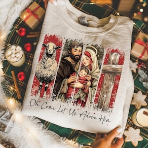 May include: A light beige crewneck sweatshirt featuring a graphic print of a sheep, the Holy Family, and a wooden cross against a red background with snowflake accents. The text "Oh Come Let Us Adore Him" is printed in black script below.