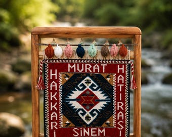 May include: A rectangular woven tapestry featuring a central geometric diamond pattern in red, white, blue, and brown, framed by the text MURAT HAKKARI HATIRASI SINEM. The tapestry hangs from a wooden frame with small colorful yarn balls on top.