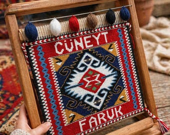 May include: A small, framed tapestry with a geometric design in red, blue, yellow, and white. The tapestry is held in a wooden frame and features the names "CUNEYT" and "FARUK". Decorative yarn balls in various colors are attached to the top.