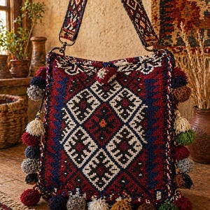 May include: A woven shoulder bag with a geometric pattern in red, blue, black, and white. The bag features a patterned strap and is adorned with colorful pom-poms along the bottom and sides. The bag is a bohemian style.