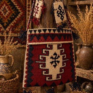 May include: A woven shoulder bag with a geometric design in red, black, and cream. The bag has a long strap and is adorned with tassels. Two smaller matching pouches hang from the wooden post. The background features a rug with similar patterns.