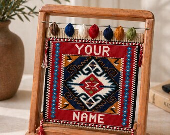 May include: A rectangular woven tapestry featuring a geometric pattern in red, blue, white, and yellow, with the text "YOUR NAME" embroidered in white. The tapestry hangs from a metal rod on a wooden frame, accented by small colorful yarn tassels.
