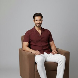 May include: A maroon short-sleeved shirt with a textured pattern, paired with white pants and brown leather shoes. The outfit is worn by a person seated in a brown armchair against a neutral background. The shirt is buttoned at the top.