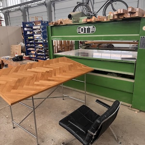 May include: A workshop scene featuring a wooden desk with a herringbone pattern, a black leather chair, and a large green machine. A bicycle rests on top of the machine, and the room has a high ceiling.