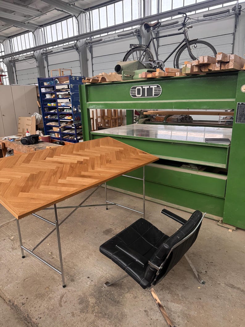 May include: A workshop scene featuring a wooden desk with a herringbone pattern, a black leather chair, and a large green machine. A bicycle rests on top of the machine, and the room has a high ceiling.
