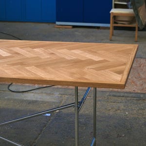 May include: A rectangular wooden table with a herringbone pattern on the tabletop. The table has a light brown finish and is supported by chrome legs. The table's dimensions are approximately 60 inches long and 30 inches wide.