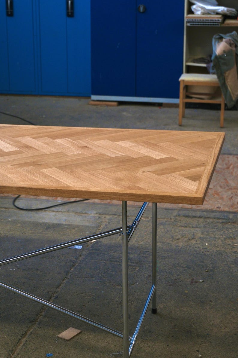 May include: A rectangular wooden table with a herringbone pattern on the tabletop. The table has a light brown finish and is supported by chrome legs. The table's dimensions are approximately 60 inches long and 30 inches wide.