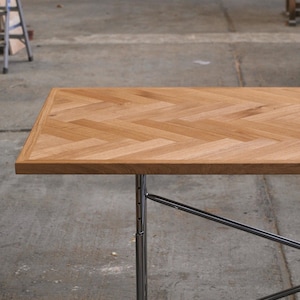 Op de afbeelding: Een rechthoekige houten tafel met een visgraatpatroon op het tafelblad. De tafel heeft een lichtbruine afwerking en wordt ondersteund door chromen poten. De tafel is ongeveer 122 cm lang.