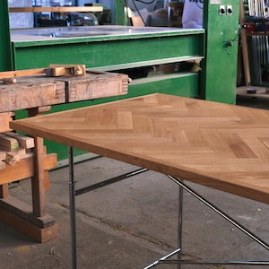 May include: A light brown wooden table with a herringbone pattern top and chrome legs. The table is in a workshop setting with green machinery and a wooden workbench in the background.