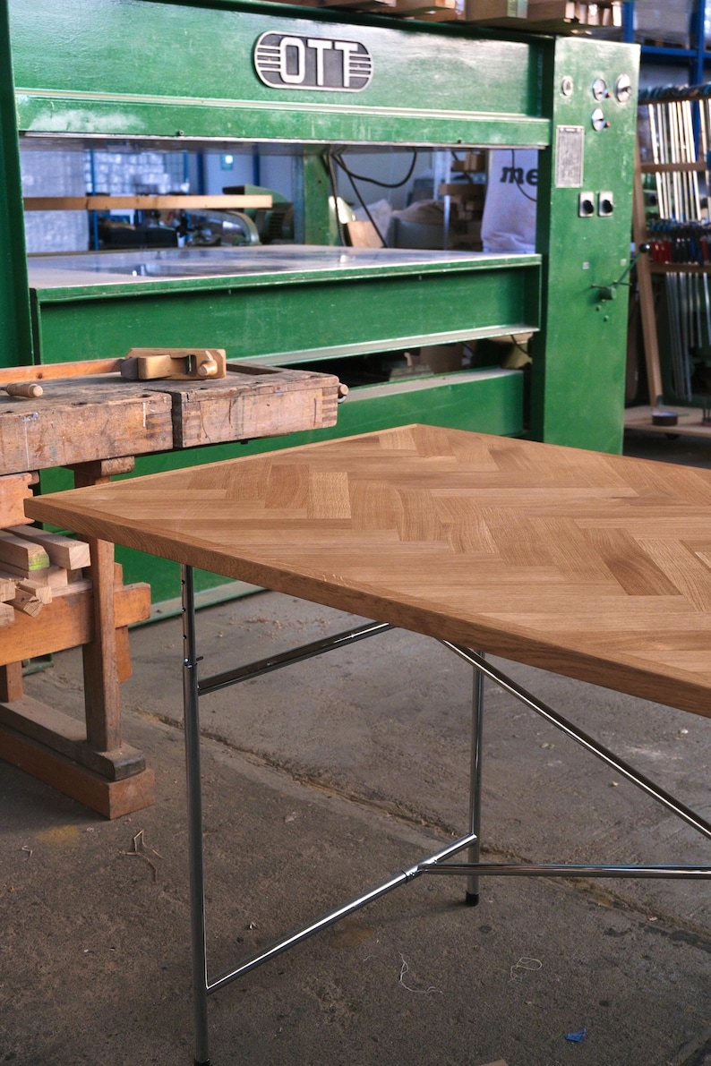 May include: A light brown wooden table with a herringbone pattern top and chrome legs. The table is in a workshop setting with green machinery and a wooden workbench in the background.