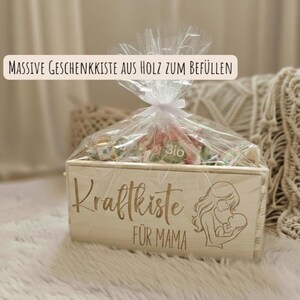 Könnte beinhalten: Geschenkbox aus hellem Holz mit der Aufschrift "Kraftkiste für Mama" und einer Illustration von Mutter und Kind. Die Box ist mit verschiedenen Artikeln gefüllt und in Zellophan verpackt.