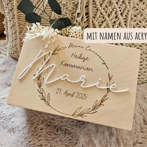 Könnte beinhalten: Ein Holzschild mit dem Namen "Marie" in weißer Schrift, umgeben von einem floralen Kranz. Das Schild enthält den Text "Meine Erinnerungen Heilige Kommunion 27. April 2025" und "MIT NAMEN AUS ACRYL". Der Hintergrund ist eine weiche, neutrale Farbe.