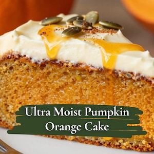 May include: A slice of Ultra Moist Pumpkin Orange Cake on a white plate. The cake is orange with white frosting, drizzled with a golden glaze, and topped with pumpkin seeds. The text "Ultra Moist Pumpkin Orange Cake" is visible.