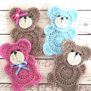 May include: Four handmade crochet teddy bear appliques in shades of brown, pink, and blue. Each bear has a white muzzle and black eyes. The pink bear has a blue bow, and the brown bear has a pink bow with a pearl.