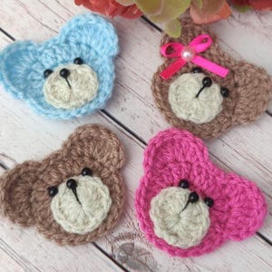 May include: Four handmade crochet bear appliques in blue, brown, and pink. Each bear has a cream-colored face, black bead eyes, and a black stitched nose. One brown bear has a pink bow with a pearl.