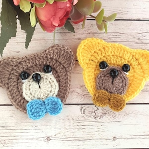 May include: Two crocheted teddy bear head appliques. One is brown with a blue bow tie, the other is yellow with a brown bow tie. Both have black button eyes and a light-colored muzzle. The appliques are on a light wood background.
