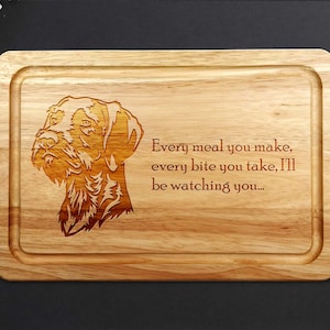 May include: A rectangular wooden chopping board featuring a carved dog's head and the quote: "Every meal you make, every bite you take, I'll be watching you...". The board has a recessed edge.