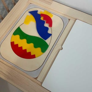May include: A colourful wooden egg puzzle, featuring red, yellow, green, and blue pieces. The puzzle sits within a light-coloured wooden frame, with a white surface to the right. A child's toy.